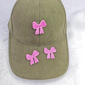 Könnte beinhalten: Olivgrüne Cord-Baseballkappe mit drei rosa Schleifenapplikationen. Die Kappe hat einen gebogenen Schirm und einen Knopf oben. Das Wort "Positivity" ist im Hintergrund sichtbar.