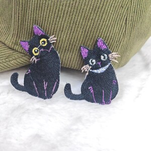 May include: Two embroidered patches of black cats with purple ears and accents. The cats have yellow eyes and tan whiskers. One cat is sitting, and the other is in a similar pose. The patches are on a white surface.