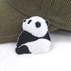 Op de afbeelding: Een wit en zwart panda beer patch met geborduurde details. De panda heeft zwarte ogen, oren en vlekken op zijn lichaam. De patch ligt op een wit oppervlak, met een groene corduroy hoed op de achtergrond.