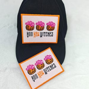 May include: Black baseball cap with a rectangular patch featuring three orange jack-o'-lanterns wearing pink witch hats. The patch has the text "BOO HA! WITCHES" in orange and white. A second patch is also visible.