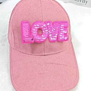 Può includere: Cappellino da baseball rosa con la scritta "LOVE" a lettere di paillettes sul davanti. Il cappellino è realizzato in tessuto strutturato e ha una visiera curva. Lo sfondo è bianco.