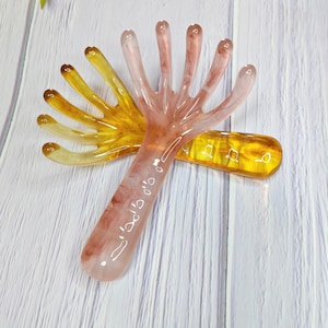 Peut inclure: Deux outils de massage en forme de main. L'un est de couleur ambre translucide, et l'autre est rose translucide avec des marbrures marron. Les outils sont posés sur une surface en bois blanc. Les outils sont conçus pour masser le cuir chevelu.