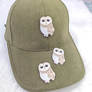 Op de afbeelding: Olijfgroene corduroy baseballpet met drie geborduurde uilenpatches. De uilen zijn wit en beige met zwarte ogen. De pet heeft een gebogen klep en een knoop aan de bovenkant.