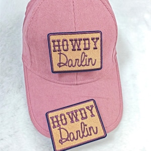 Puede incluir: Gorra de béisbol de pana rosa polvoriento con visera curva. La gorra presenta un parche rectangular con las palabras "HOWDY Darlin" bordadas en cursiva. También es visible un parche a juego.