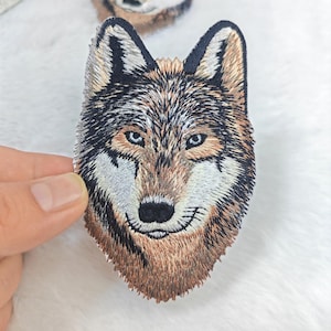 Puede incluir: Parche bordado de lobo con una representación detallada de la cabeza de un lobo. El parche muestra un diseño realista con hilo marrón, negro y blanco, capturando el pelaje y los rasgos faciales del lobo. El lobo tiene ojos azules y una nariz negra.
