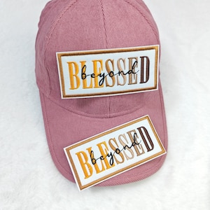 May include: A dusty rose corduroy baseball cap with the embroidered text "BLESSED beyond" in a rectangular patch. A smaller matching patch is below the cap. The text is in a mix of yellow, brown, and black.