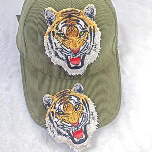 Pode incluir: Boné de beisebol de veludo cotelê verde-oliva com dois patches bordados de cabeça de tigre. As cabeças de tigre têm detalhes em laranja, preto e branco com bocas abertas e línguas vermelhas. O boné tem uma aba curva.