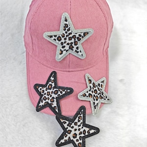 May include: Pink corduroy baseball cap with three star patches. The stars have a leopard print design with silver and black borders. The cap is a solid pink color.