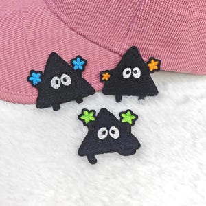 Könnte beinhalten: Drei schwarze, dreieckige Stickaufnäher mit weißen ovalen Augen und kleinen bunten Sternakzenten in Blau, Orange und Limettengrün, platziert auf einem strukturierten rosa Stoff mit einem sichtbaren, geschwungenen Rand oben.