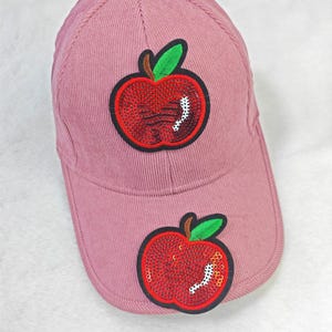 Könnte beinhalten: Staubrosa Baseballkappe mit Cordstruktur. Die Kappe hat zwei gestickte rote Apfelaufnäher mit grünen Blättern und einem weißen Akzent. Die Äpfel sind mit roten Pailletten verziert. Die Kappe hat einen gebogenen Schirm.