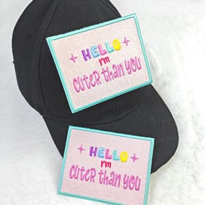Könnte beinhalten: Schwarze Baseballkappe mit zwei gestickten Aufnähern. Jeder Aufnäher hat einen rosa Hintergrund, einen hellblauen Rand und den Text "HELLO I'M Cuter than you" in bunten Fäden.