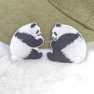 Peut inclure: Deux écussons brodés représentant des pandas. Chaque écusson présente un panda noir et blanc en position assise. Les écussons sont posés sur une surface blanche et duveteuse, avec un chapeau vert en velours côtelé en arrière-plan.