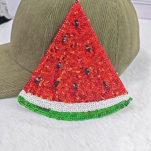 Pode incluir: Uma fatia de melancia vermelha com lantejoulas com detalhes de sementes pretas, acabamento branco e verde, repousando sobre um boné de beisebol de veludo cotelê verde. Um acessório atraente.