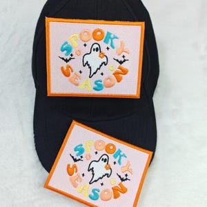May include: Black baseball cap with a rectangular patch featuring the text "SPOOKY SEASON" around a ghost illustration. The patch has a pink background with orange, blue, and yellow lettering and an orange border. Bats are also included in the design.
