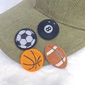Op de afbeelding: Een groene corduroy baseball cap met vier geborduurde sportpatches: een voetbal, een achtbal, een basketbal en een American football. De patches zijn in zwart, wit, oranje en bruin.