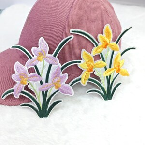 Pode incluir: Boné de beisebol rosa empoeirado com dois patches florais bordados. Um patch apresenta flores roxas e amarelas, o outro tem flores amarelas. Os patches têm caules e folhas verdes.