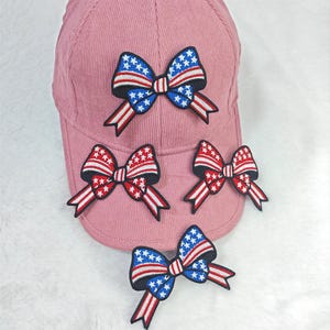Pode incluir: Um boné de beisebol rosa empoeirado adornado com quatro patches de laço bordados. Os laços apresentam um design patriótico com listras vermelhas e brancas, campos azuis de estrelas e contornos pretos. O boné é feito de tecido tipo veludo cotelê.