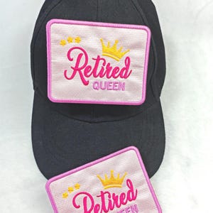 Könnte beinhalten: Schwarze Baseballkappe mit einem rosa bestickten Aufnäher mit der Aufschrift "Retired Queen" in Schreibschrift. Der Aufnäher zeigt eine goldene Krone und Sterne. Ein zweiter Aufnäher ist darunter sichtbar.