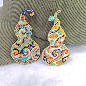 May include: Two embroidered gourd-shaped patches. One patch features a black background with blue, red, and gold swirls. The other patch has a white background with orange, blue, and gold swirls. Both patches have a gold border and are placed on a green hat.
