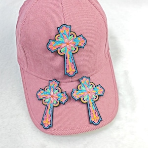 May include: A dusty rose baseball cap with three embroidered crosses. Each cross features a colorful floral design with blue, pink, yellow, and green accents. The cap has a curved brim and a structured crown.