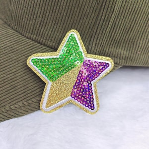 May include: A sequined star patch in green, gold, and purple, with a white border, is attached to a green corduroy hat. The star is divided into sections of green sequins, gold glitter, and purple sequins.