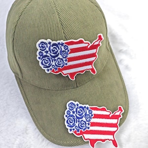 May include: An olive green corduroy baseball cap with two embroidered patches. Each patch is a map of the United States with a floral design in blue and red stripes, resembling the American flag.