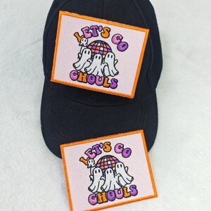 May include: Black baseball cap with a rectangular patch featuring the text "Let's Go Ghouls" in purple and orange. The patch includes a graphic of three white ghosts and a colorful globe on a pink background.