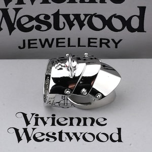 May include: A silver-toned ring with a unique, angular design. The ring features multiple geometric shapes and small, decorative studs. The words "Vivienne Westwood JEWELLERY" are visible in the background.