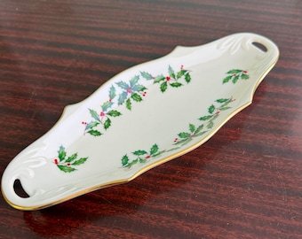 May include: An elongated, off-white porcelain serving dish with a gold-trimmed edge and a holly leaf and red berry pattern. The dish features a symmetrical, curved shape with a small, circular cutout handle on each end of the platter.