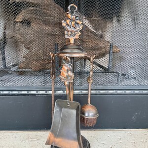 May include: A copper-toned fireplace tool set featuring a stand with a ship motif at the top. The set includes a shovel and a brush, both with matching ship-themed handles. The metal finish has a dark, aged patina, and the tools hang from a central circular frame supported by a round base.