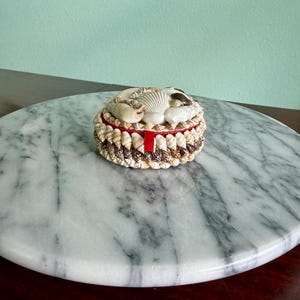 May include: A round, decorative box adorned with various seashells. The lid and sides are embellished with a variety of shells, and a red ribbon accents the box's midsection. The box is set on a marble-like surface.