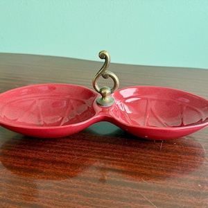 May include: A red ceramic divided serving dish with a brass handle. The dish has two oval compartments connected by a central stem. The handle is ornate and curved. The dish is on a wooden surface.