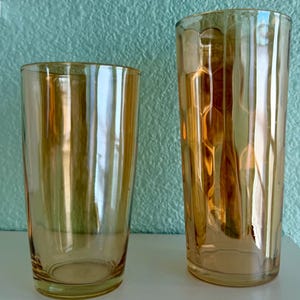 May include: Two amber-colored glass tumblers. The tumbler on the left is a simple, straight-sided design. The tumbler on the right has a textured, hexagonal pattern. Both glasses are empty and reflect the light.