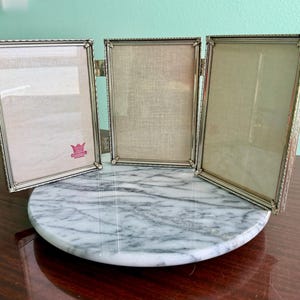 May include: A silver-toned, three-panel picture frame with ornate detailing. The frame is hinged and stands on a round, marble-patterned base. Each panel has a rectangular opening for photos. A pink logo is visible on the left panel.