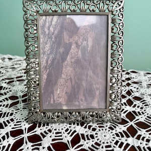 May include: Ornate silver-colored picture frame with a detailed, scrolling design. The frame holds a faded image. The frame is displayed on a dark wooden surface covered with a white lace doily.