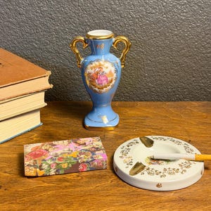 May include: A blue ceramic vase with gold handles and a painted scene, a floral-decorated ashtray with a cigarette, and a floral-patterned box are on a wooden surface. The vase is approximately 5 inches tall.