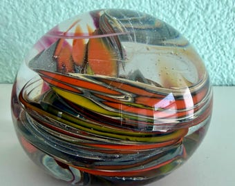 May include: A clear glass paperweight with colorful swirls. The interior features layers of red, orange, yellow, and black glass, creating a dynamic, abstract design. The spherical shape is smooth and polished, reflecting light and adding visual interest to any desk or shelf.
