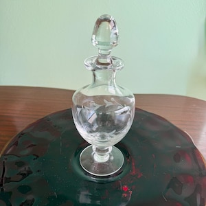 May include: A clear glass perfume bottle with a decorative stopper. The bottle has a rounded body with etched floral designs and sits on a circular base. The stopper is a rounded shape. The bottle is displayed on a dark red glass plate.