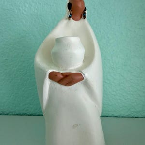 May include: A white ceramic figurine of a Native American woman in a flowing robe, holding a small white vase. The woman's face is brown, with black hair and blue earrings. The figurine is set against a light blue background.