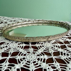 May include: Oval silver-toned vanity tray with a mirrored surface. The tray has an ornate, openwork border. The background is a white lace doily on a dark wooden surface, with a light green wall in the background.