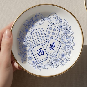 May include: A round white ceramic dish with a gold-toned rim, featuring a blue line-art illustration of three mahjong tiles surrounded by a floral wreath and a bow. The tiles display bamboo, dot, and Chinese character designs in blue ink.