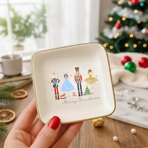 May include: A square, off-white ceramic trinket dish with a gold rim. The dish features a festive illustration of Nutcracker characters and the words "Merry Christmas". The dish is held by a person.