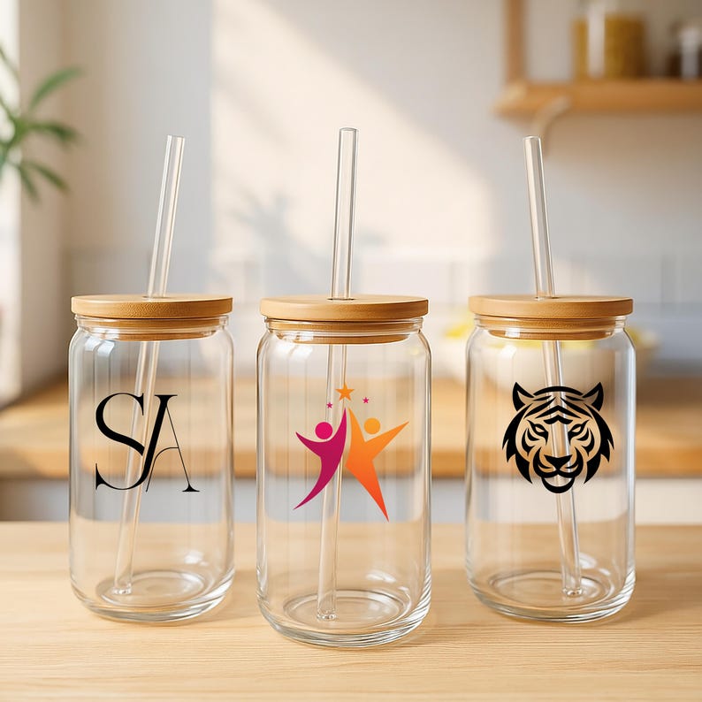 May include: Three clear glass drinking jars with bamboo lids and clear straws. Each jar has a different design: the letters "SA", a graphic of two people with stars, and a tiger head. The jars are on a wooden surface.