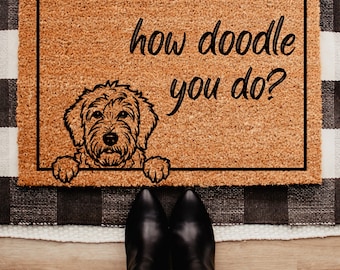 May include: A rectangular doormat with a brown coir surface and a black border. The mat features a black graphic of a dog and the phrase "how doodle you do?" in a handwritten style font. Black boots and blue jeans are visible.