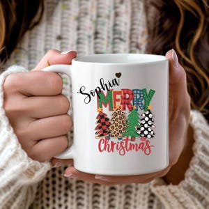Puede incluir: Taza de cerámica blanca con asa, con el nombre "Sophia" en cursiva y las palabras "Merry Christmas" en un diseño festivo. El diseño incluye cuadros escoceses, estampado de leopardo y árboles de Navidad decorados.
