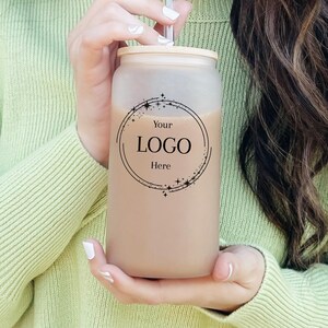 May include: A frosted glass can-shaped tumbler with a bamboo lid and a straw. The tumbler is filled with a light brown liquid. The front of the tumbler features a circular logo design with the text "Your LOGO Here".