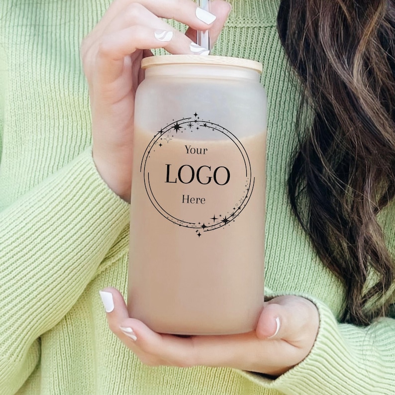 May include: A frosted glass can-shaped tumbler with a bamboo lid and a straw. The tumbler is filled with a light brown liquid. The front of the tumbler features a circular logo design with the text "Your LOGO Here".