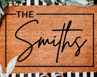 May include: A rectangular doormat with the text "THE Smiths" in a black, cursive font. The mat is a light brown color and has a textured surface. The mat is surrounded by a black border. The bottom of the image shows a pair of black shoes.