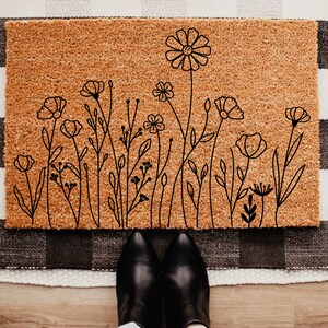 May include: A rectangular doormat with a floral design. The mat is made of a natural brown material and features a black line drawing of various flowers and foliage. The mat is placed on a black and white checkered rug.