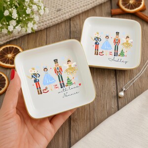 May include: Two square, white ceramic trinket dishes with gold rims. Each dish features a colorful illustration of a Nutcracker-themed design with a mouse, ballerina, and toy soldier. One dish has the name "Ashley" and the other says "with love Nannie."